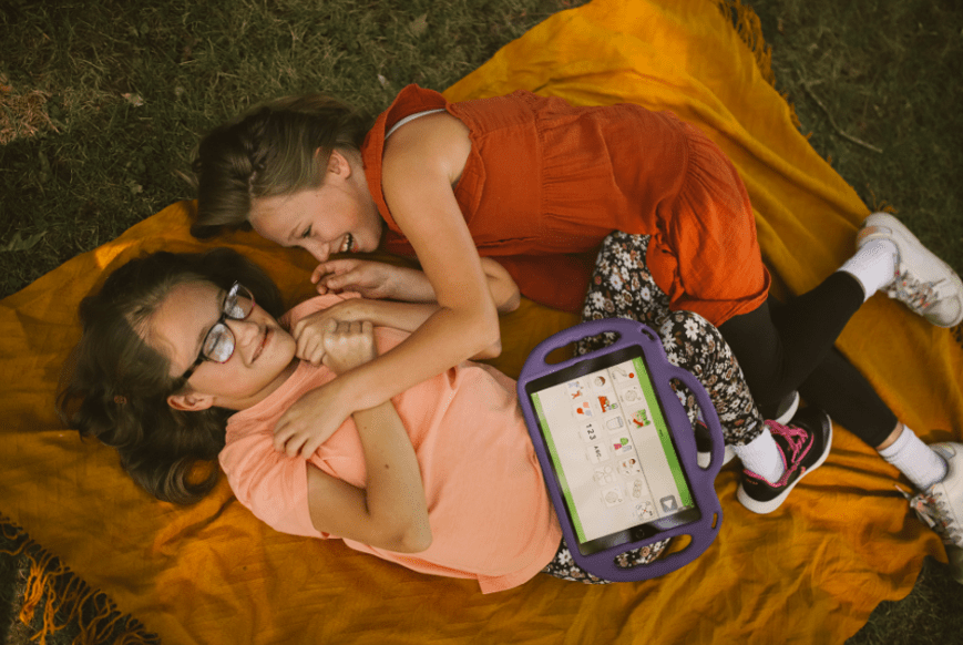 Two girls lying on a blanket, focused on a tablet, enjoying a sunny day outdoors.